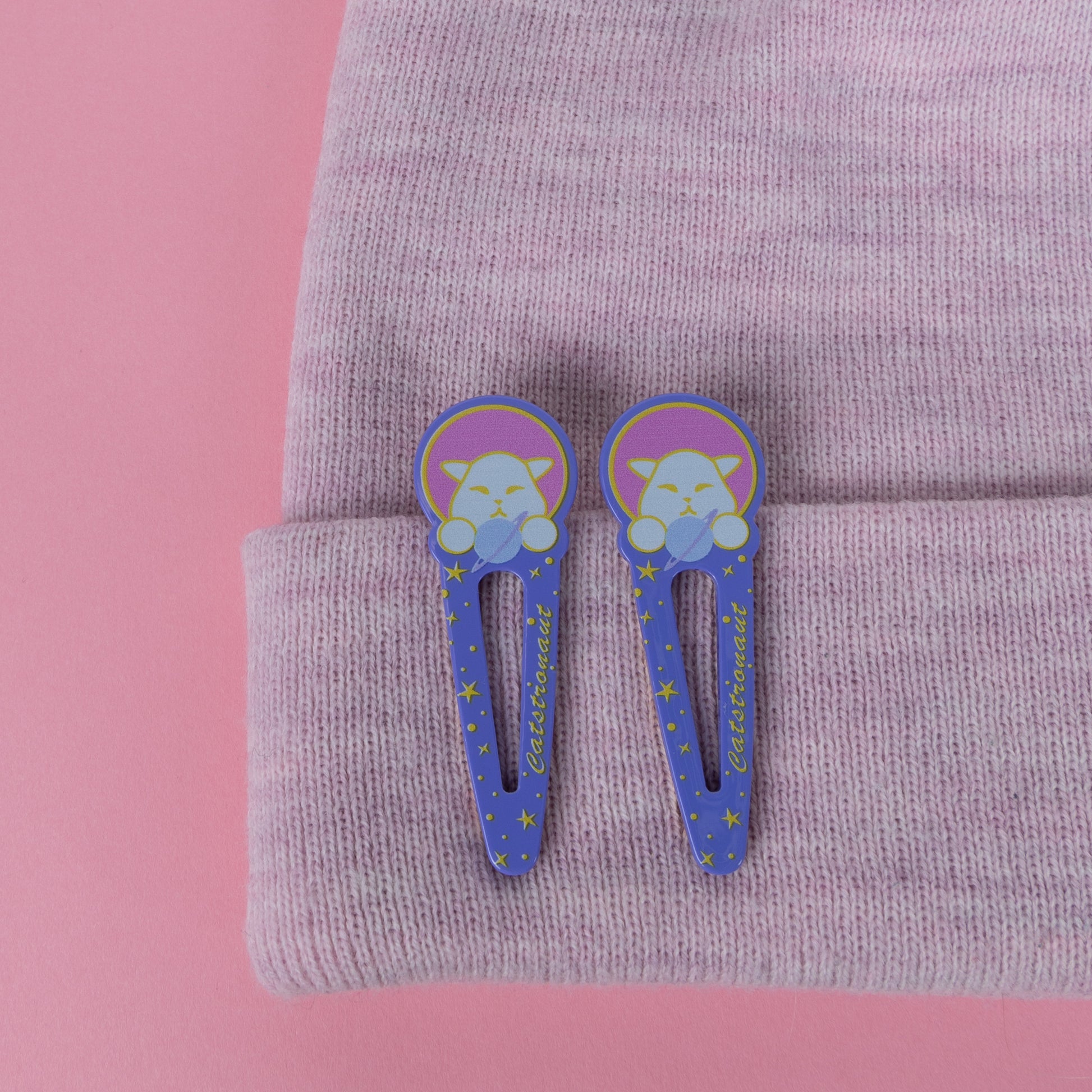 Photograph of 2 Cute Cat Astronaut (Catstronaut) acrylic pinch hair clips, featuring Catstronaut's head and paws holding a planet in the night sky; attached to the brim of a knitted pink beanie.